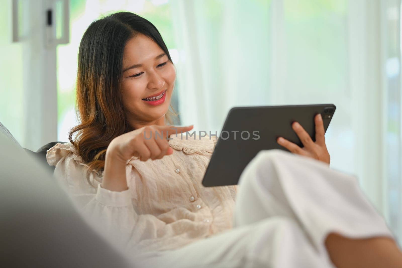 Shot of a happy young Asian woman using digital tablet while relaxing at home by prathanchorruangsak