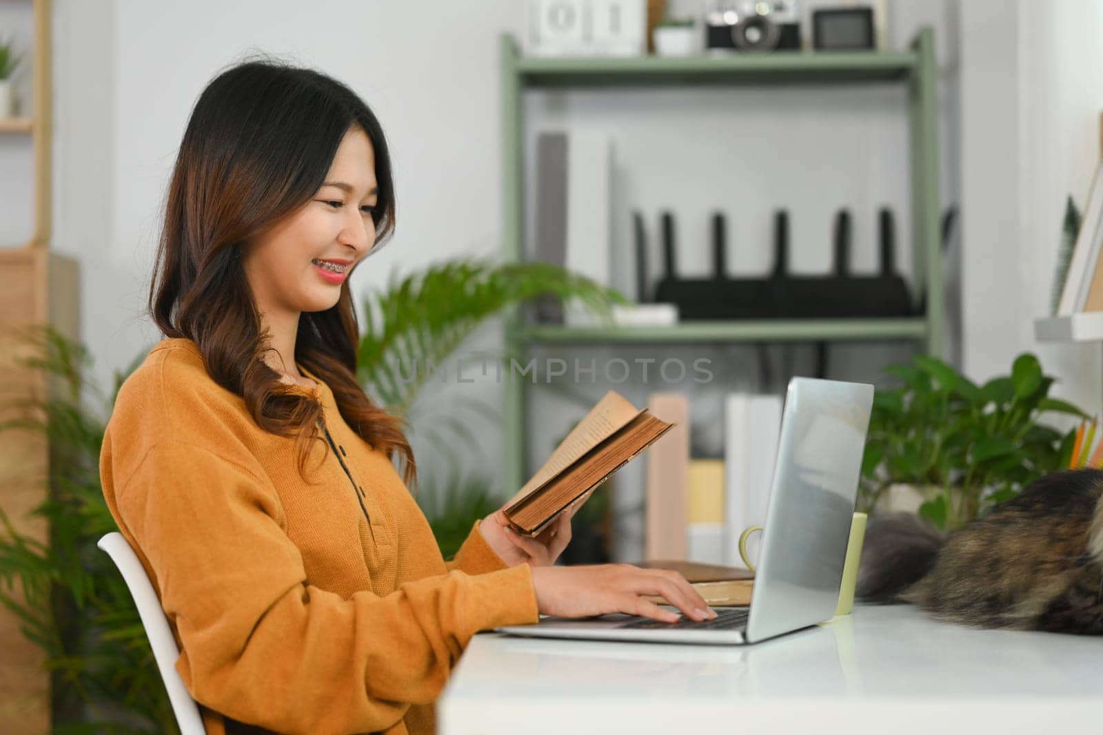 Pretty young woman holding book and working on laptop at home by prathanchorruangsak