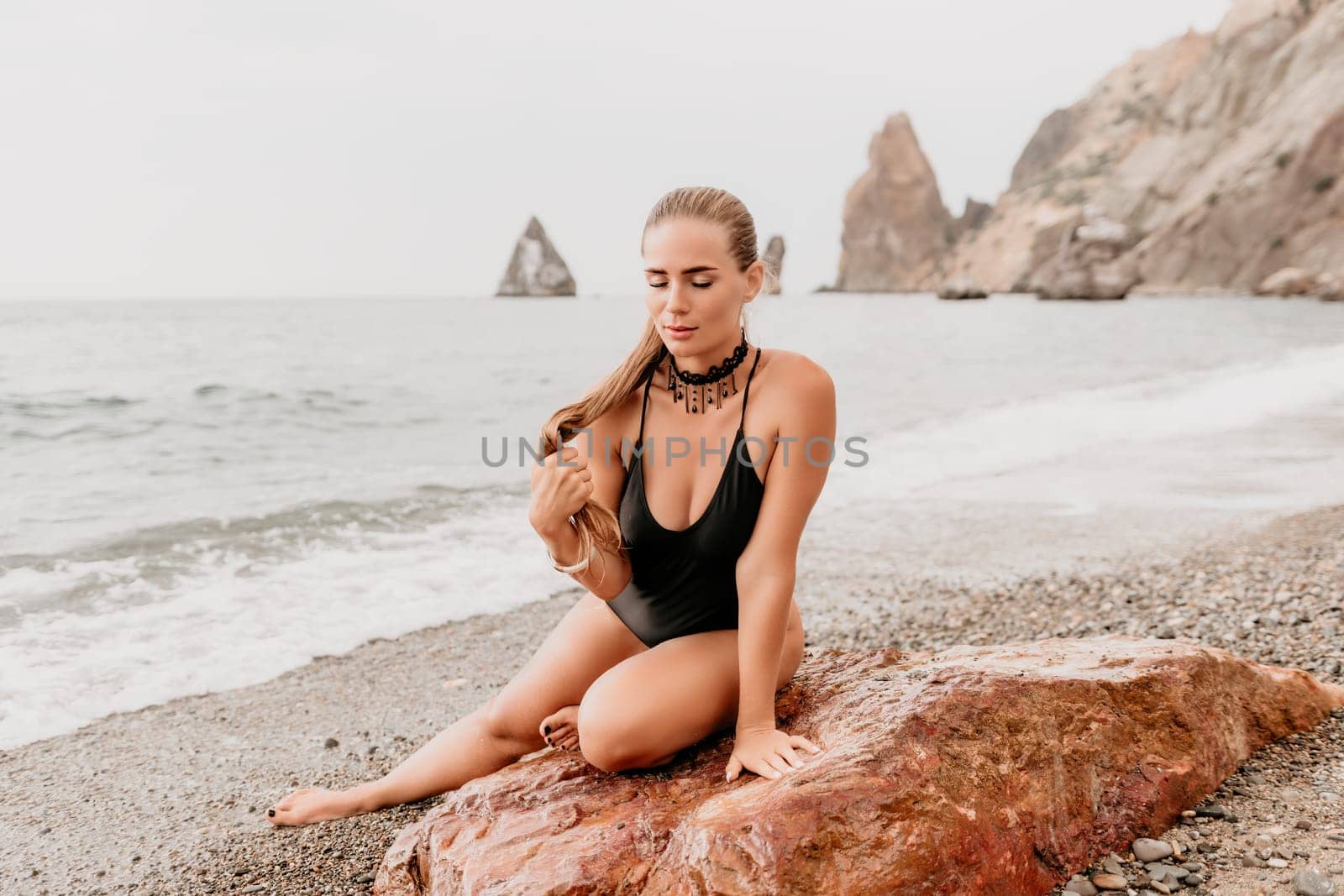 Woman travel portrait. Happy woman with long hair poses on a red volcanic rock at the beach. Close up portrait cute woman in black bikini, smiles at the camera, with the sea in the background. by panophotograph