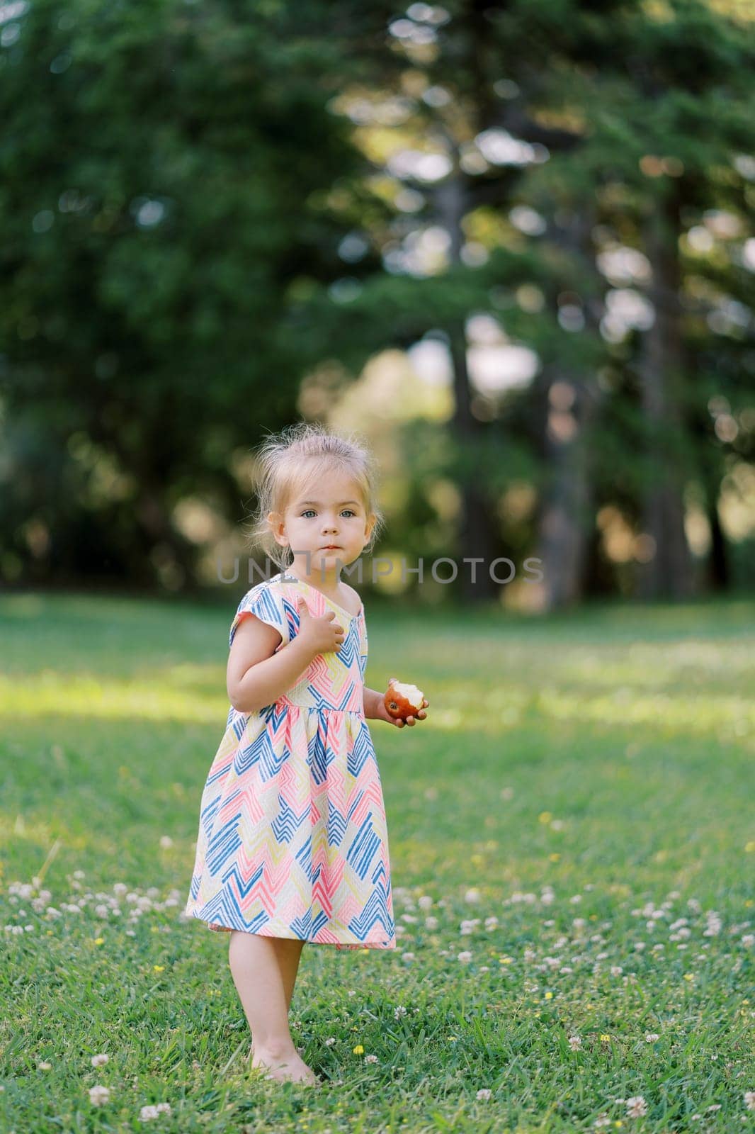 Little girl with an apple in her hand stands barefoot on a green lawn by Nadtochiy