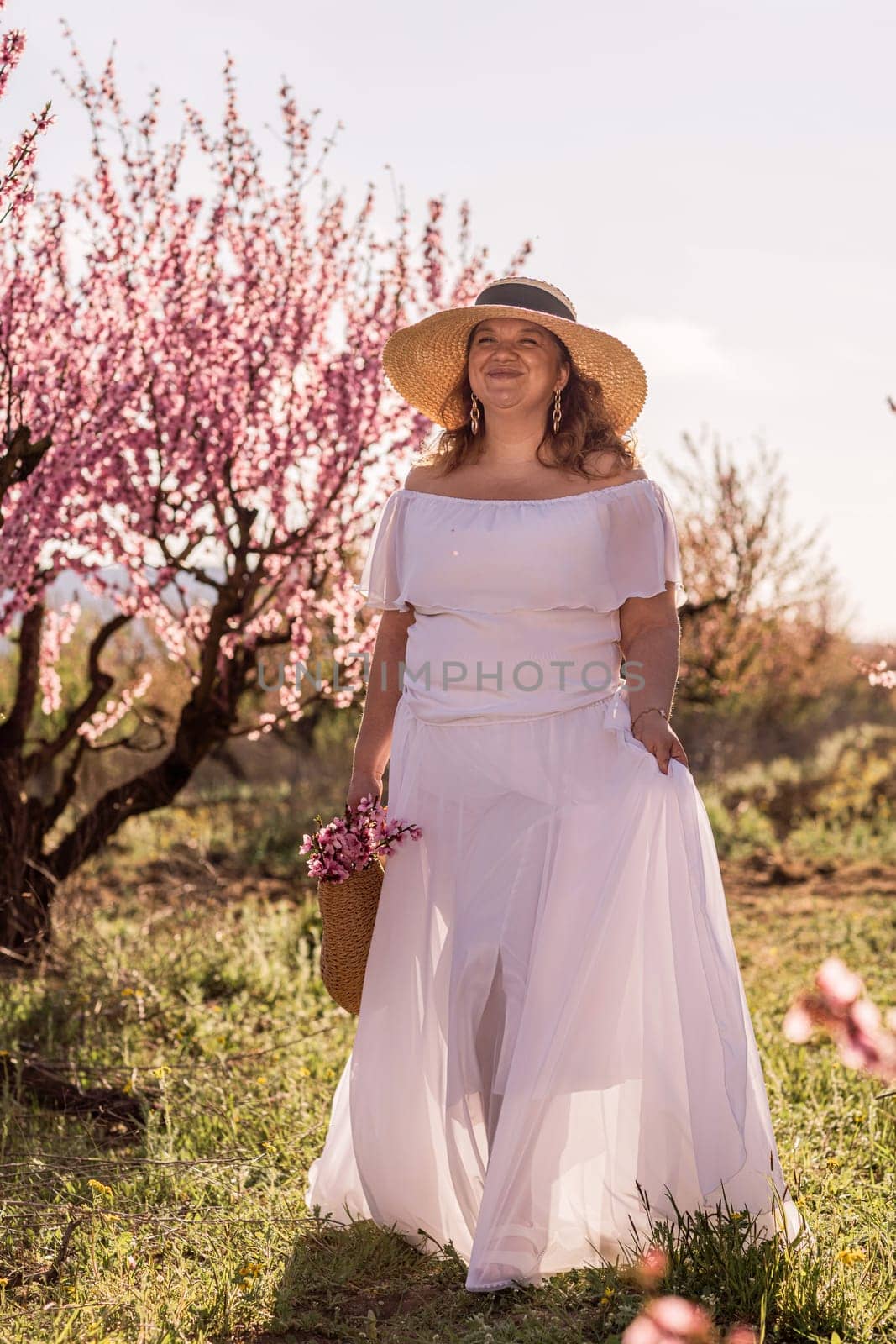 Woman blooming peach orchard. Against the backdrop of a picturesque peach orchard, a woman in a long white dress and hat enjoys a peaceful walk in the park, surrounded by the beauty of nature