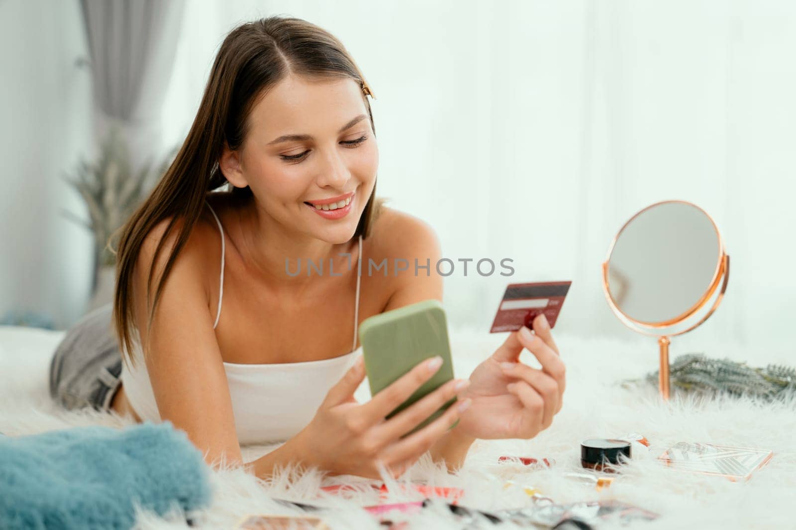 Young woman lying on the bed using online payment app and digital wallet on smartphone to pay with credit card. E commerce shopping and modern purchasing via mobile internet. Unveiling