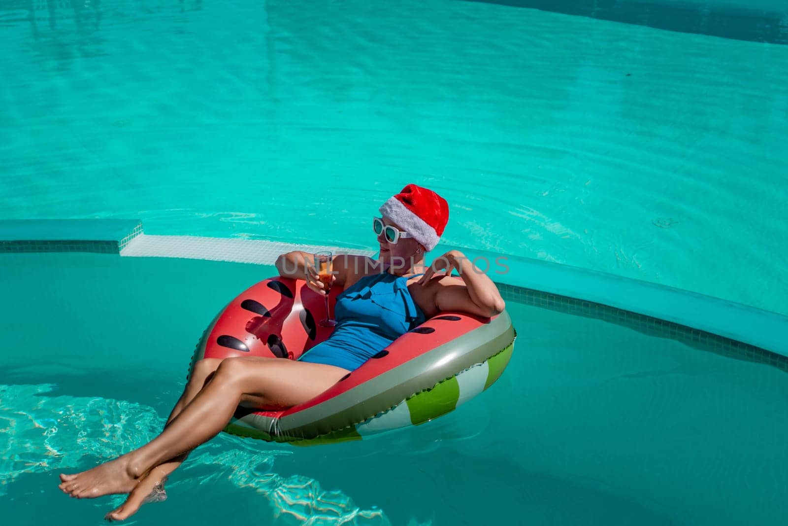 A happy woman in a blue bikini, a red and white Santa hat and sunglasses poses in the pool in an inflatable circle with a watermelon pattern, holding a glass of champagne in her hands. Christmas holidays concept