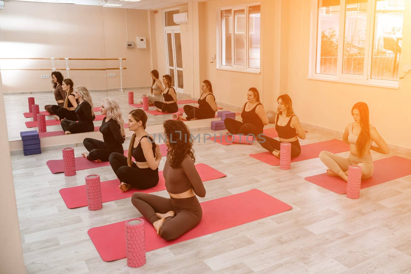 A group of six athletic women doing pilates or yoga on pink mats in front of a window in a beige loft studio interior. Teamwork, good mood and healthy lifestyle concept