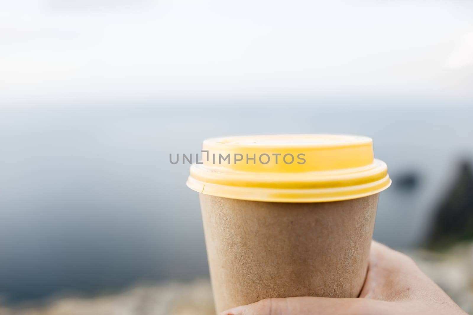 Yellow cup with lid, coffee against a backdrop of a blue sky and sea. Illustrating cup and beverage.