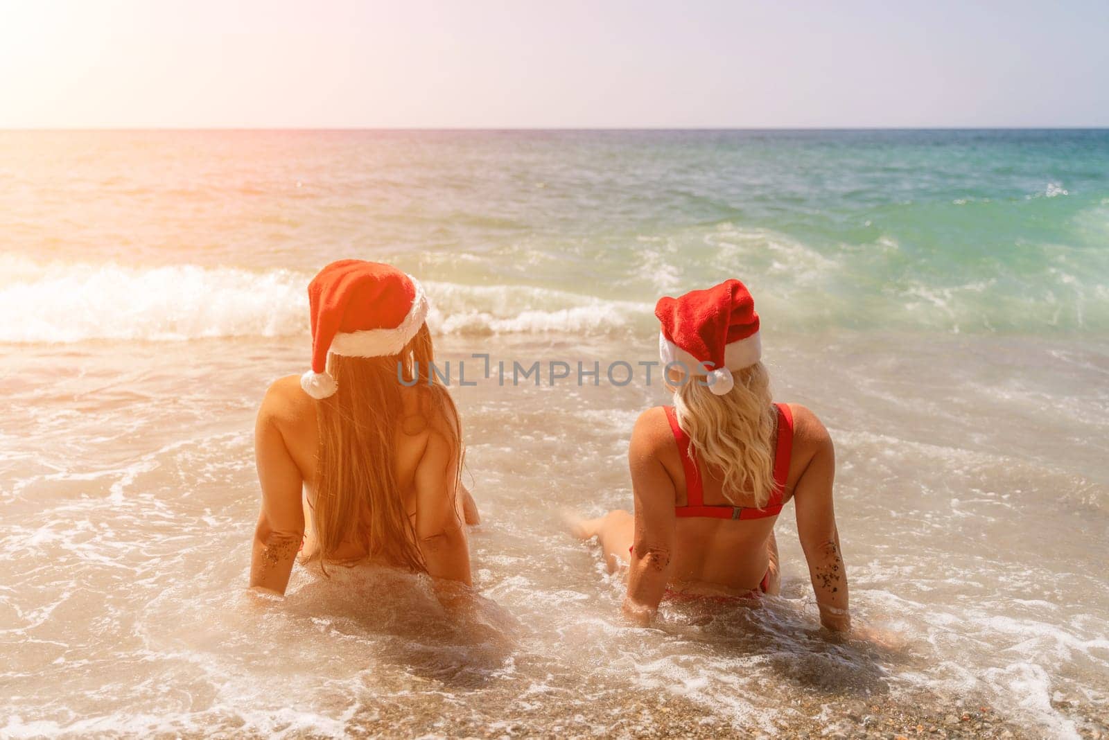 Women Santa hats ocean play. Seaside, beach daytime, enjoying beach fun. Two women in red swimsuits and Santa hats are enjoying themselves in the ocean waves. by Matiunina