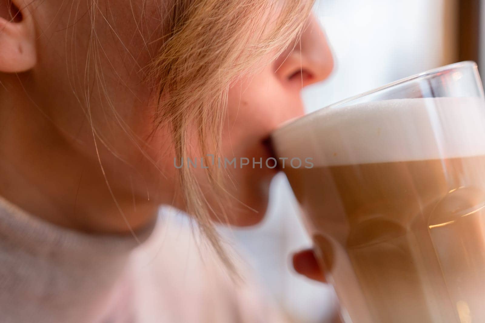 Woman with blonde hair sips cappuccino in a cafe. She is holding the glass up to her face, taking a sip of the drink