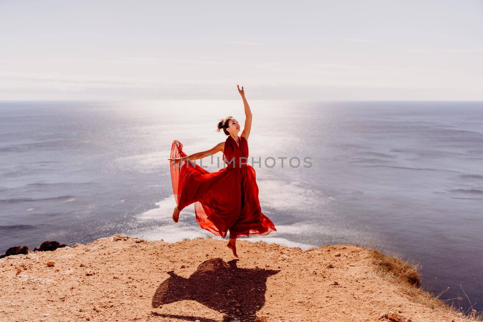 Woman red dress sea. Female dancer posing on a rocky outcrop high above the sea. Girl on the nature on blue sky background. Fashion photo