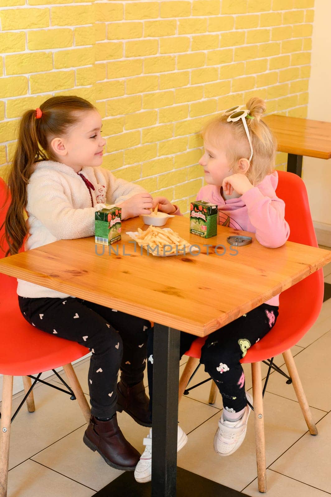 Ivano-Frankivsk, Ukraine March 26, 2023: Two little girls eat hotdogs in a cafe, junk food for children. by Niko_Cingaryuk