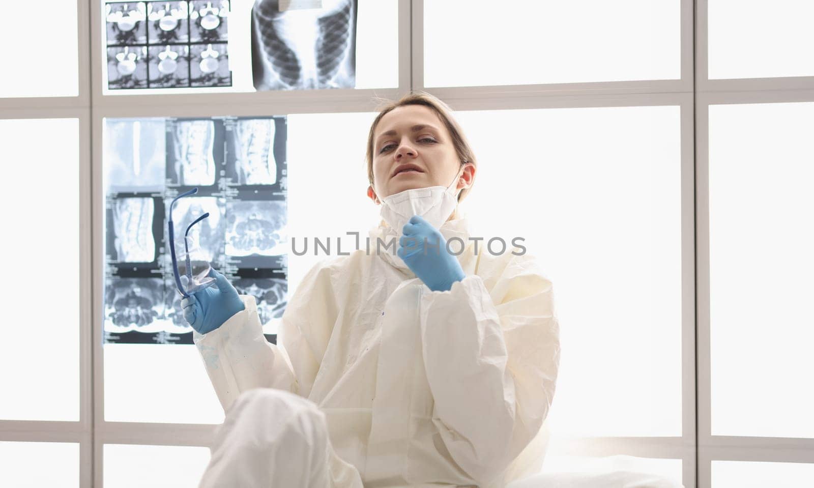 Tired doctor in protective suit sits on floor with X-rays hanging in background. Doctors who have recovered from COVID-19 concept