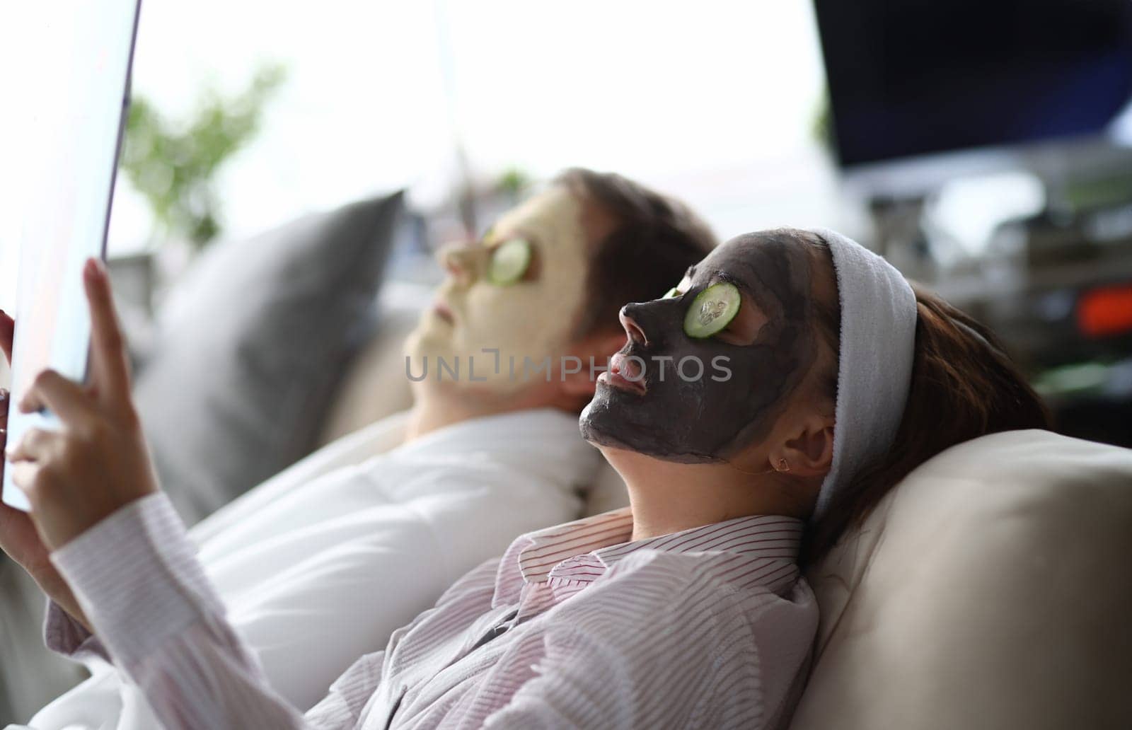 Young man and woman lying on couch with moisturizing face masks by kuprevich