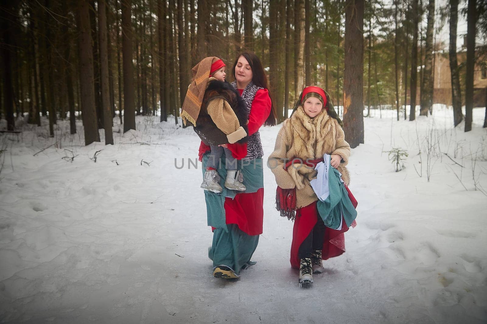 Family with mother, teenage girl, and little daughter dressed in stylized medieval peasant clothing in winter forest. Woman and her daughters pose for fairytale photoshoot in nature on a cold day