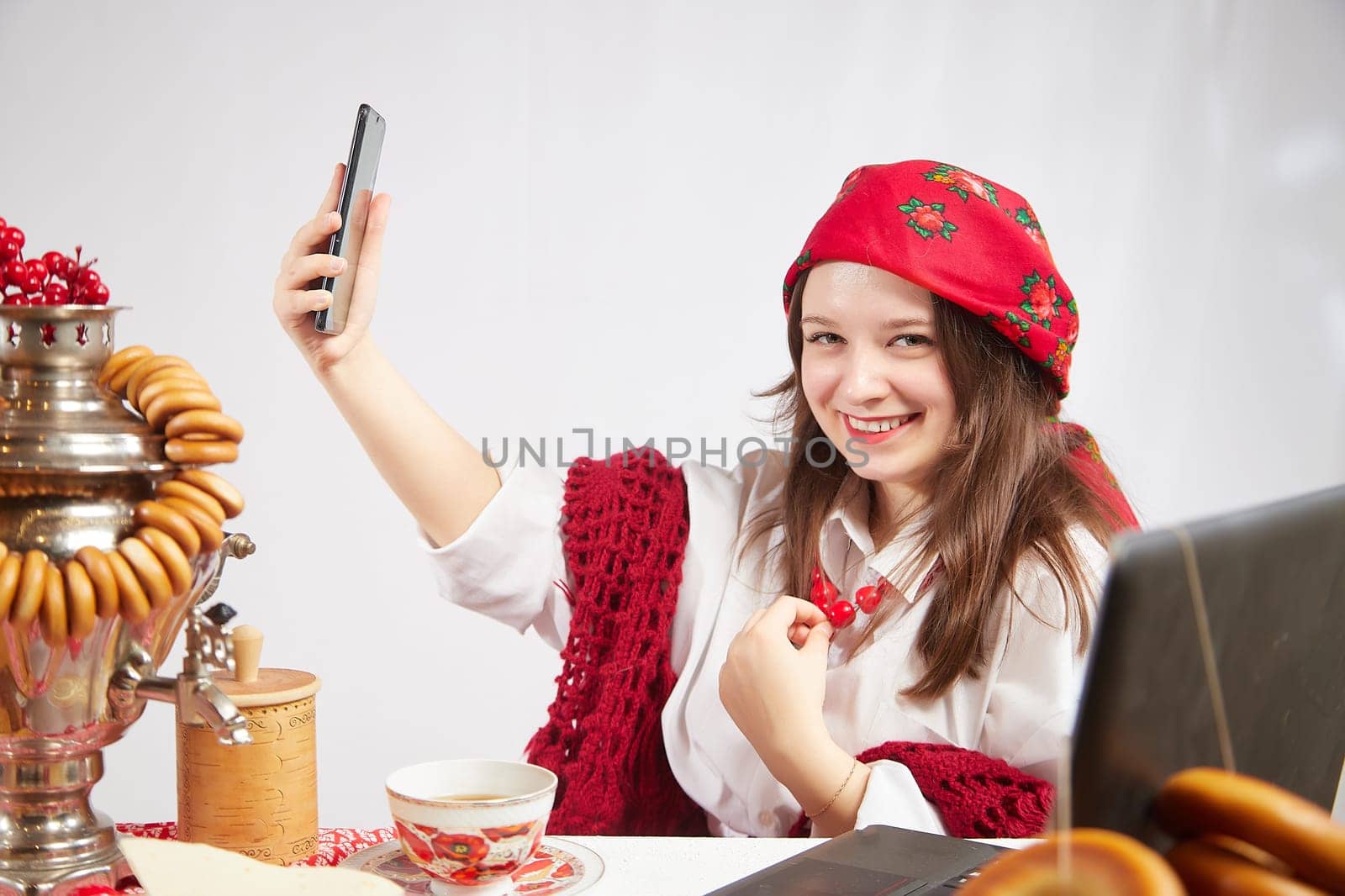 A fashionable modern girl in stylized folk clothes at table with a samovar, bagels and tea takes a selfie on the Orthodox holiday of Maslenitsa and Easter. Funny photo shoot for a young woman