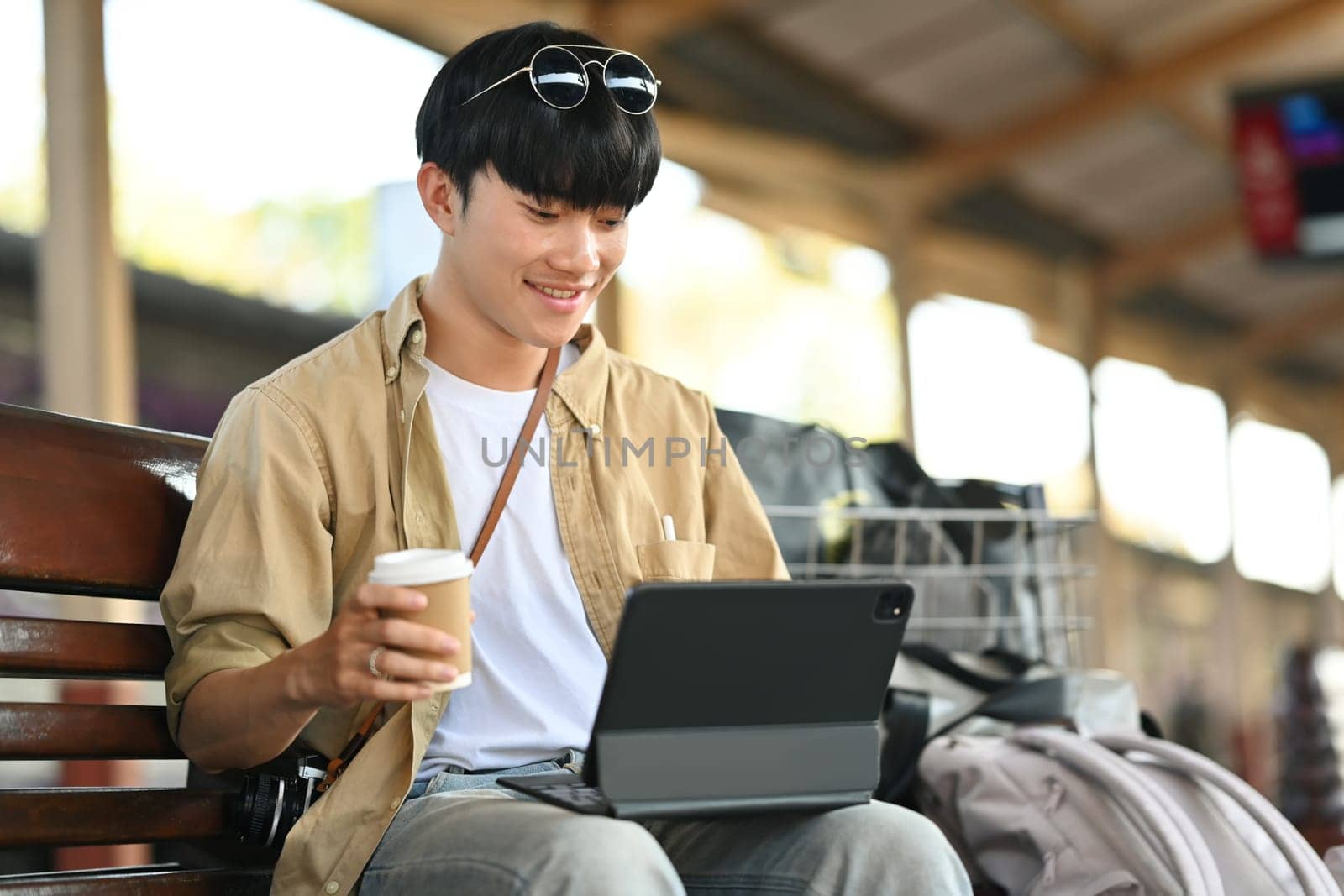 Smiling male tourist sitting with luggage at railway station and using digital tablet.