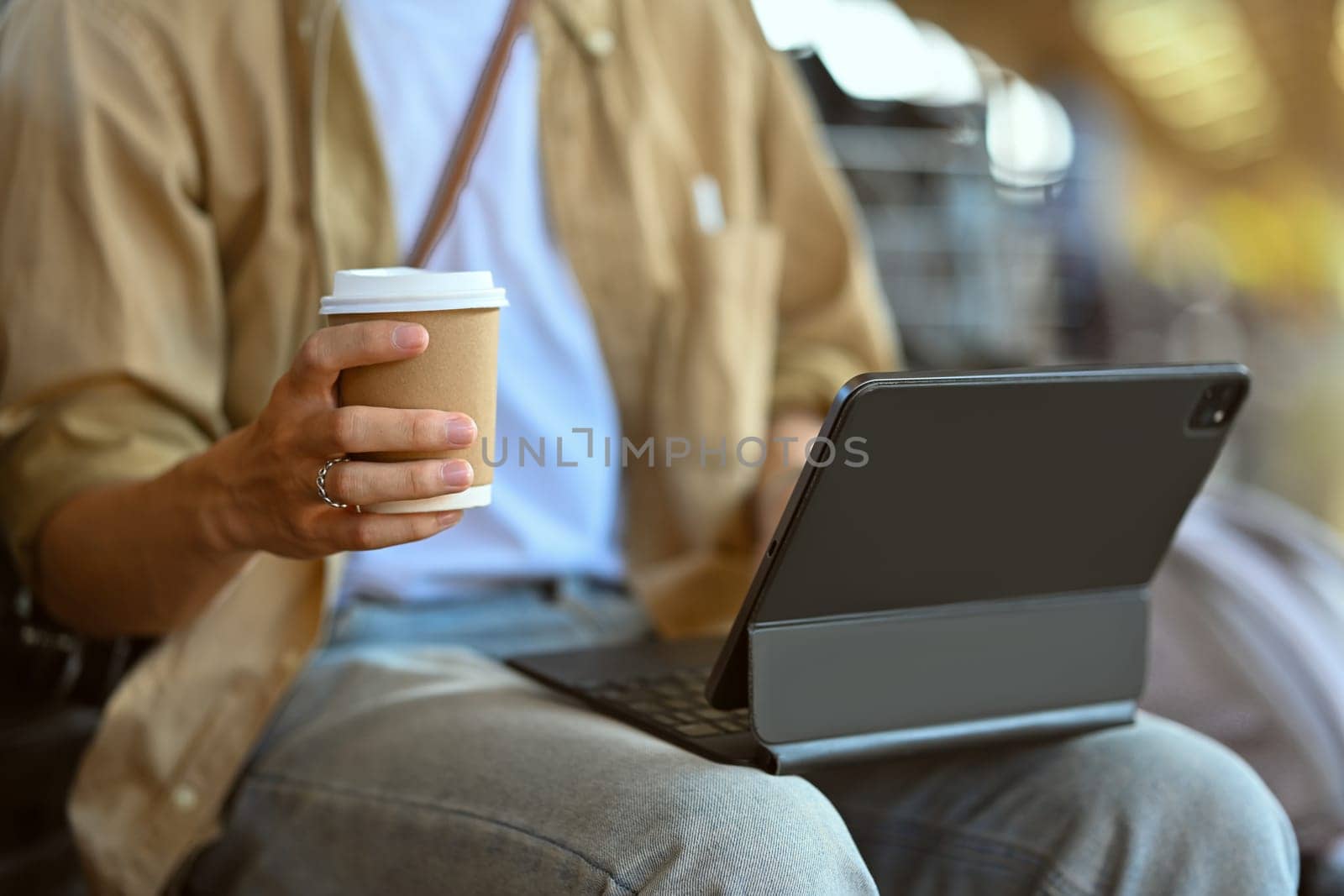 Male traveler drinking coffee and using digital tablet while waiting train at railway station by prathanchorruangsak