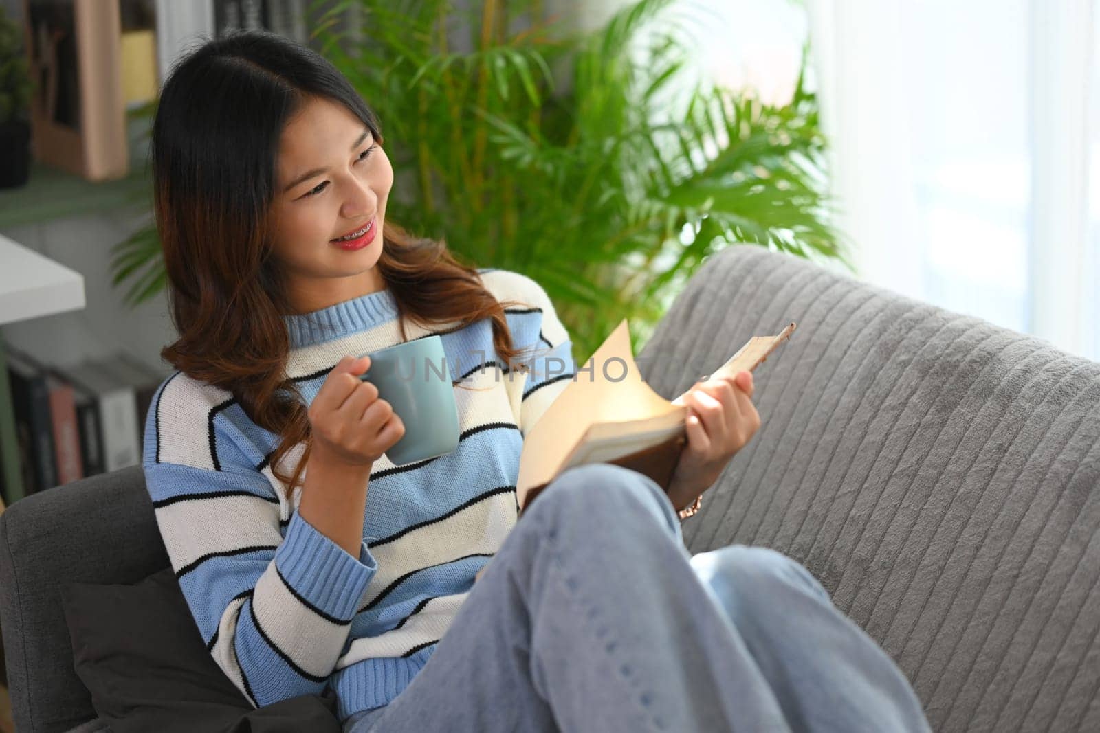 Cheerful young woman reading book and drinking hot tea on couch at home.