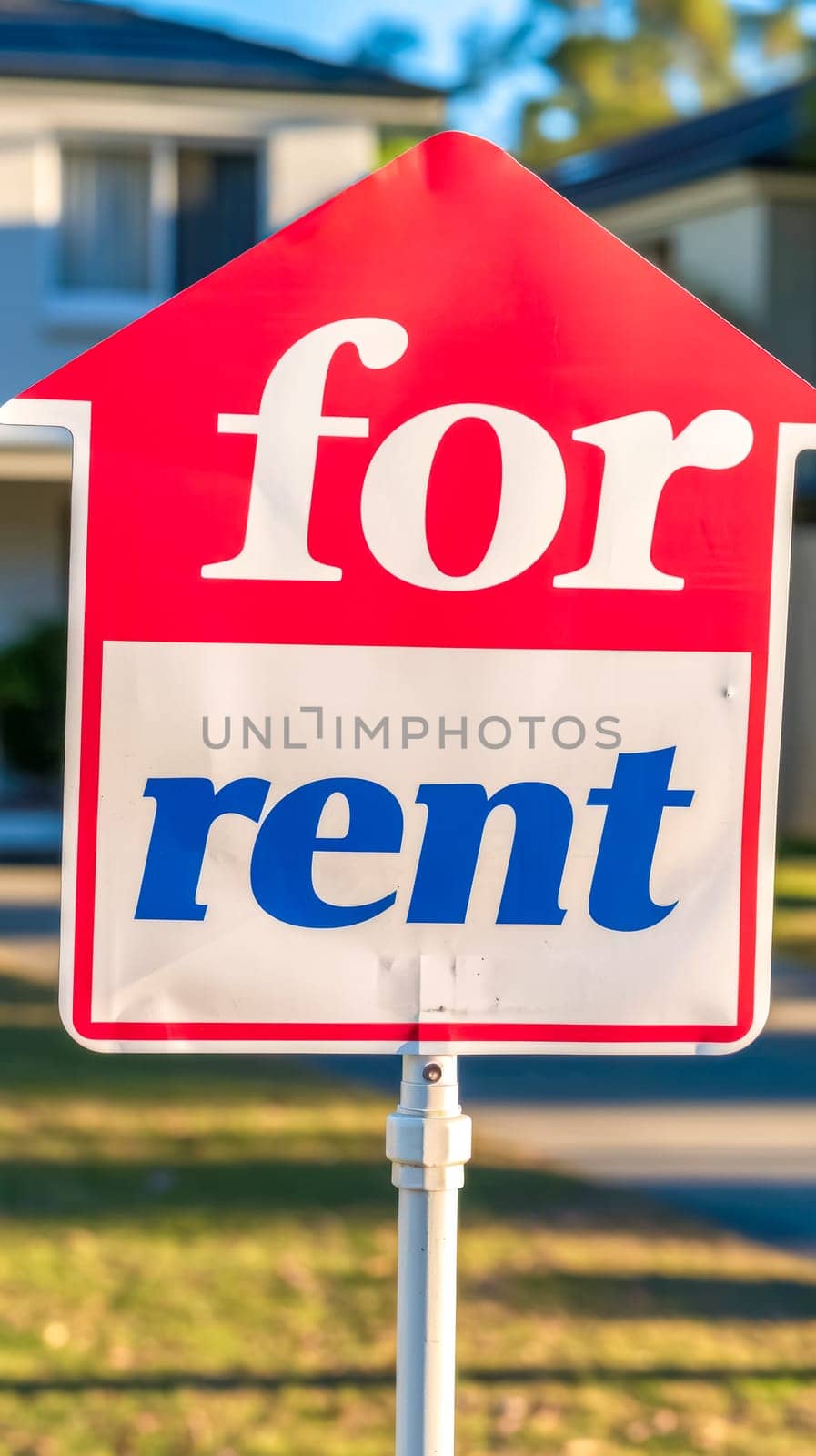 A vibrant red 'for rent' sign with bold white and blue lettering, prominently displayed on a stand with a softly blurred residential home in the background by Edophoto