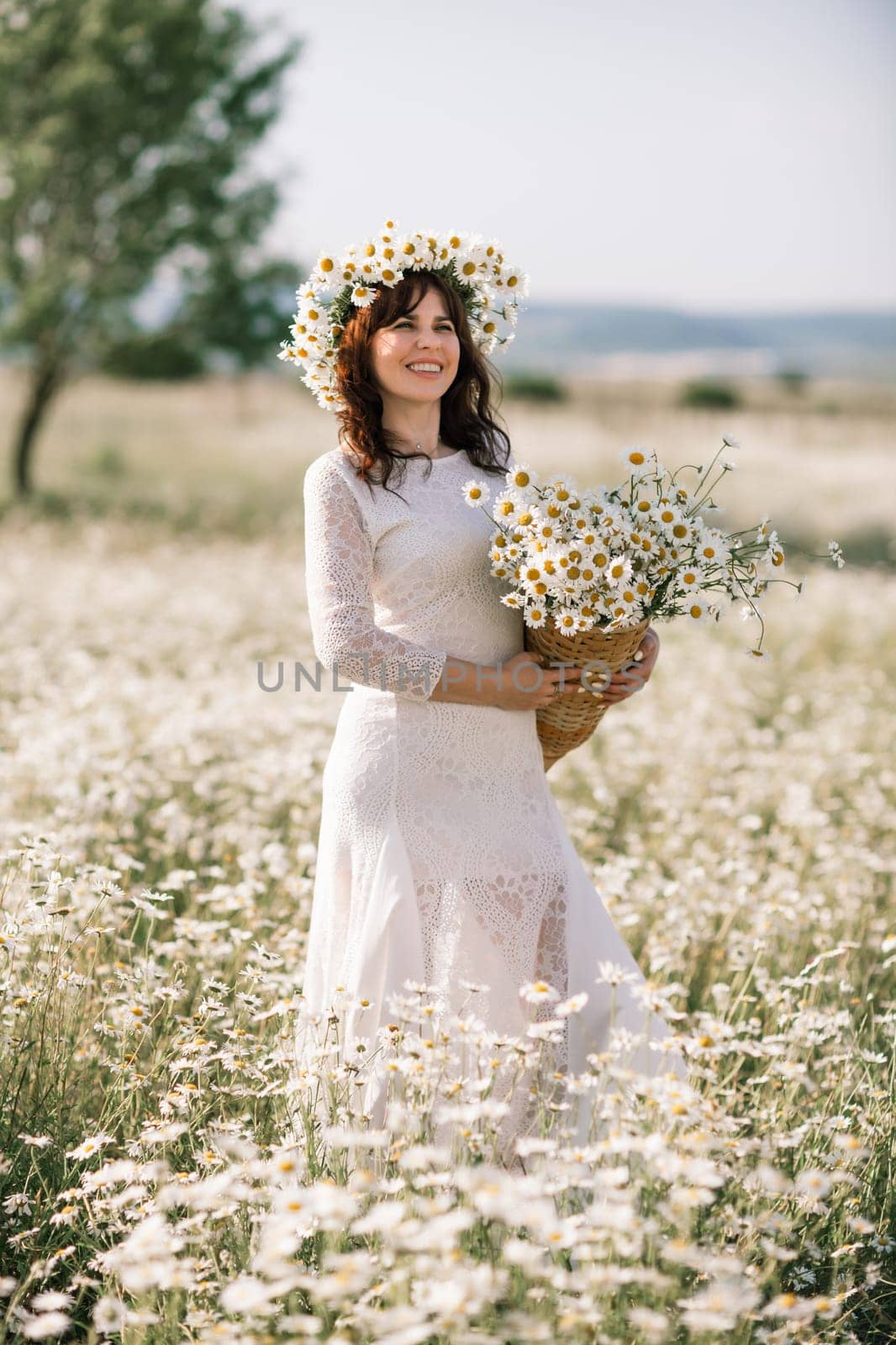 Happy woman in a field of daisies with a wreath of wildflowers on her head. woman in a white dress in a field of white flowers. Charming woman with a bouquet of daisies, tender summer photo.