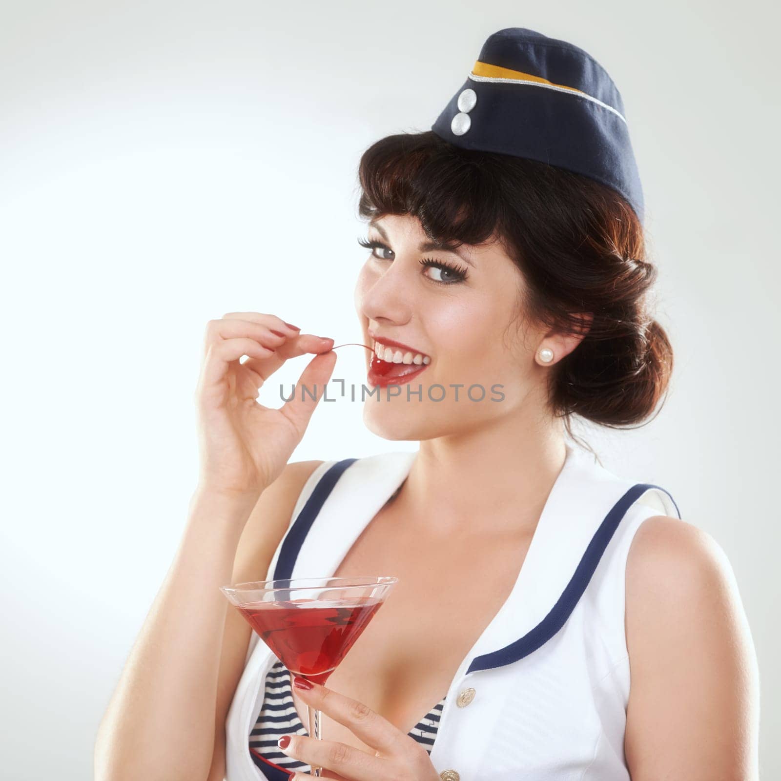 Portrait, stewardess and happy woman with alcohol to drink in studio isolated on white background. Face, martini cocktail glass and air hostess eating cherry, travel or vintage pin up girl on journey by YuriArcurs