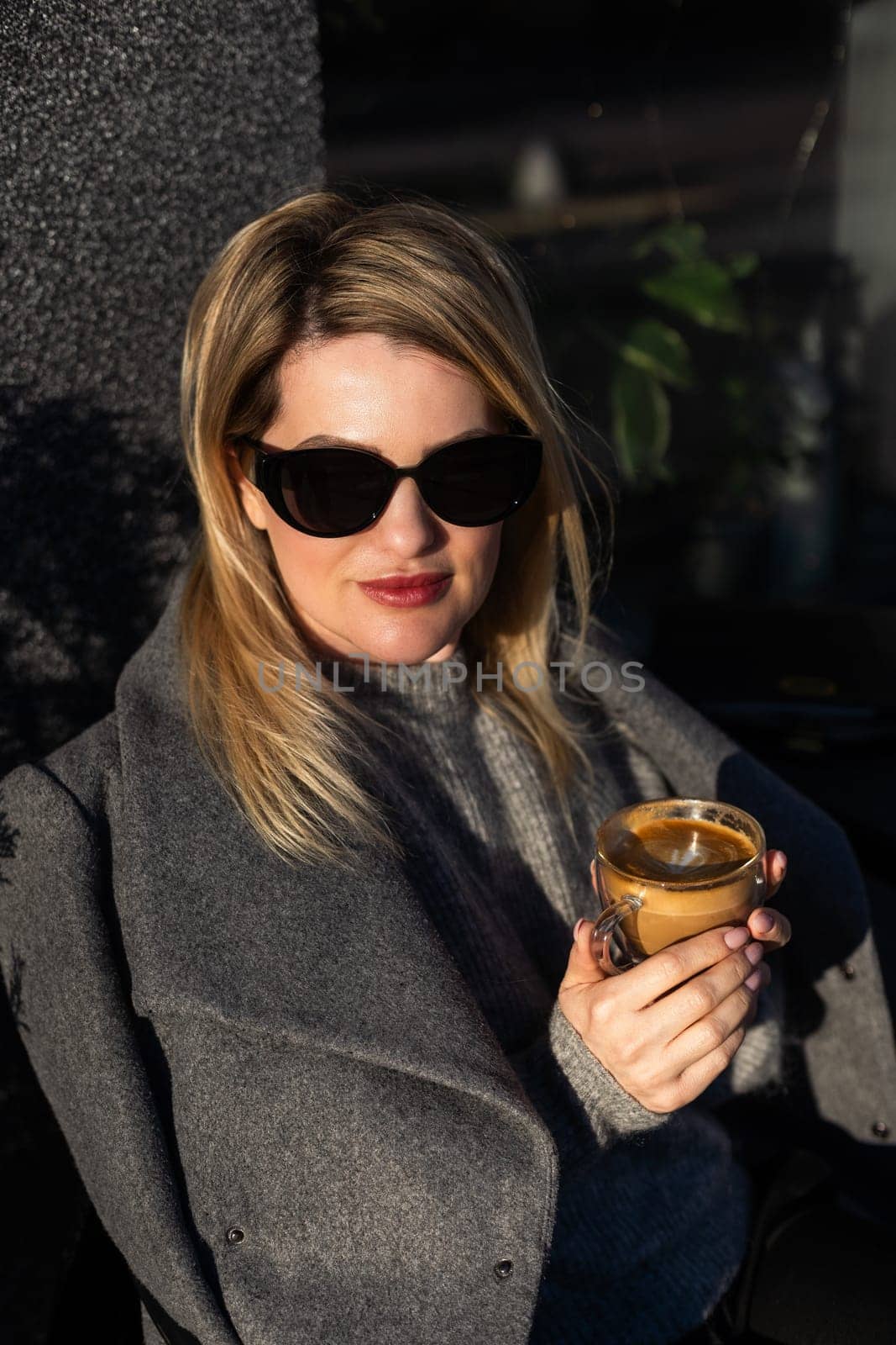 Portrait of tanned lady with cup of latte on foreground. Smiling woman in sunglasses during breakfast in cafe. High quality photo