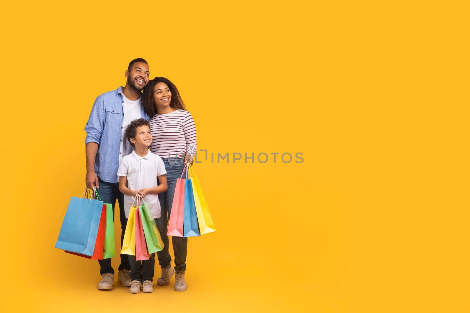Cheerful black family of three with colorful shopping bags in hands looking aside at copy space, happy african american mother, father and son enjoying seasonal sales, posing on yellow background