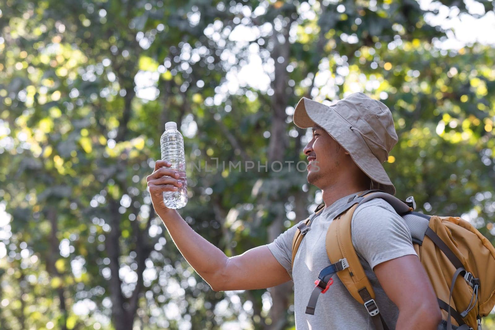 Asian male traveler carrying a large backpack drinks from a bottle while resting during a hiking trip. by wichayada