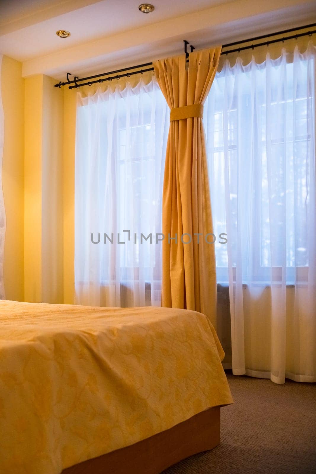 Luxury interior of bedroom with big window with white curtains. Comfortable bed with sheets and yellow pillows in modern style.