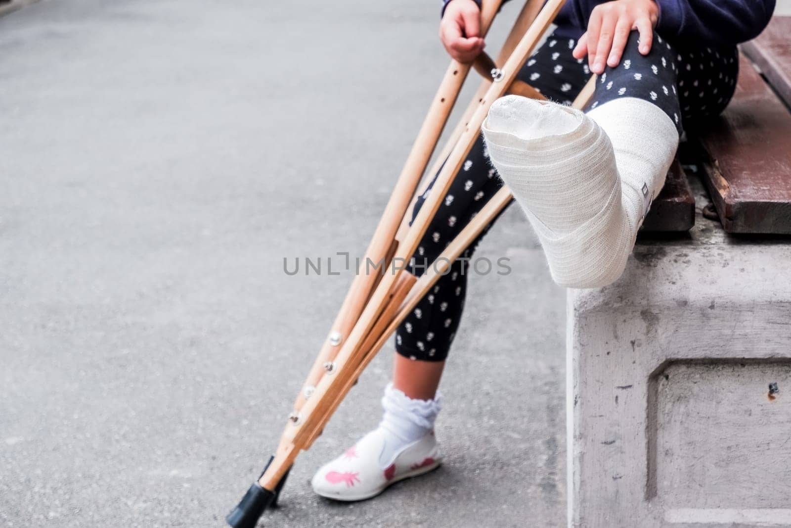 unrecognizable girl sits on the street on a bench with a broken leg and crutches.disabled girl with crutches.an accident while jumping on a trampoline. An ankle fracture. by YuliaYaspe1979