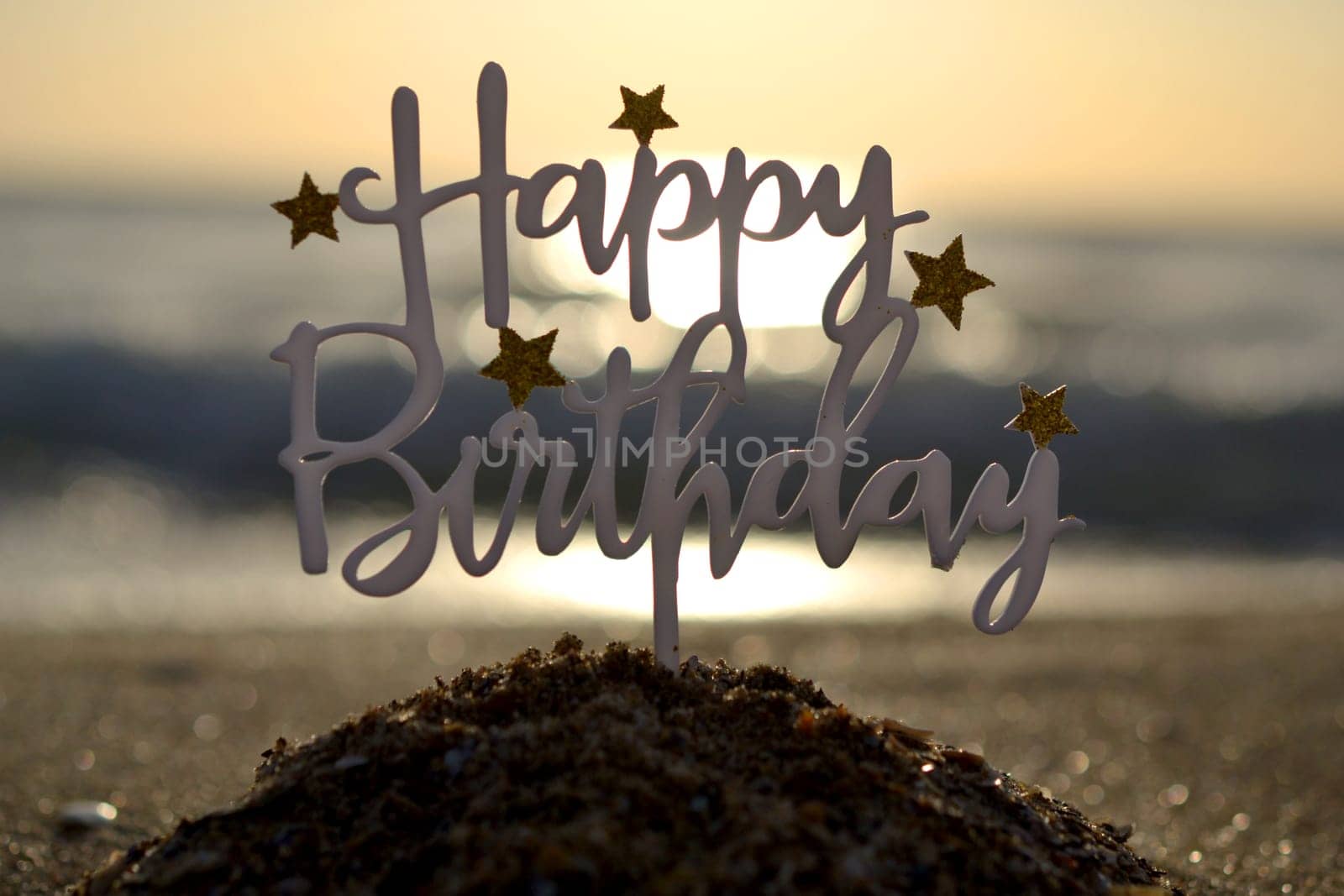 Stick in shape of letters Happy Birthday with stars at dawn and sunset on background of sea waves on sand seashore. Word Happy birthday in sandy beach on sunny summer day. Concept Happy Birthday