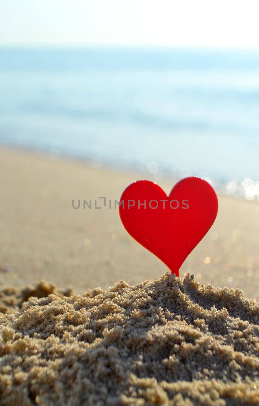 Plastic stick in shape of red heart in sand on sandy beach of sea shore on background of sea waves on sunny summer day close-up. Concept Love relationship romance amorousness amour St. Valentines Day