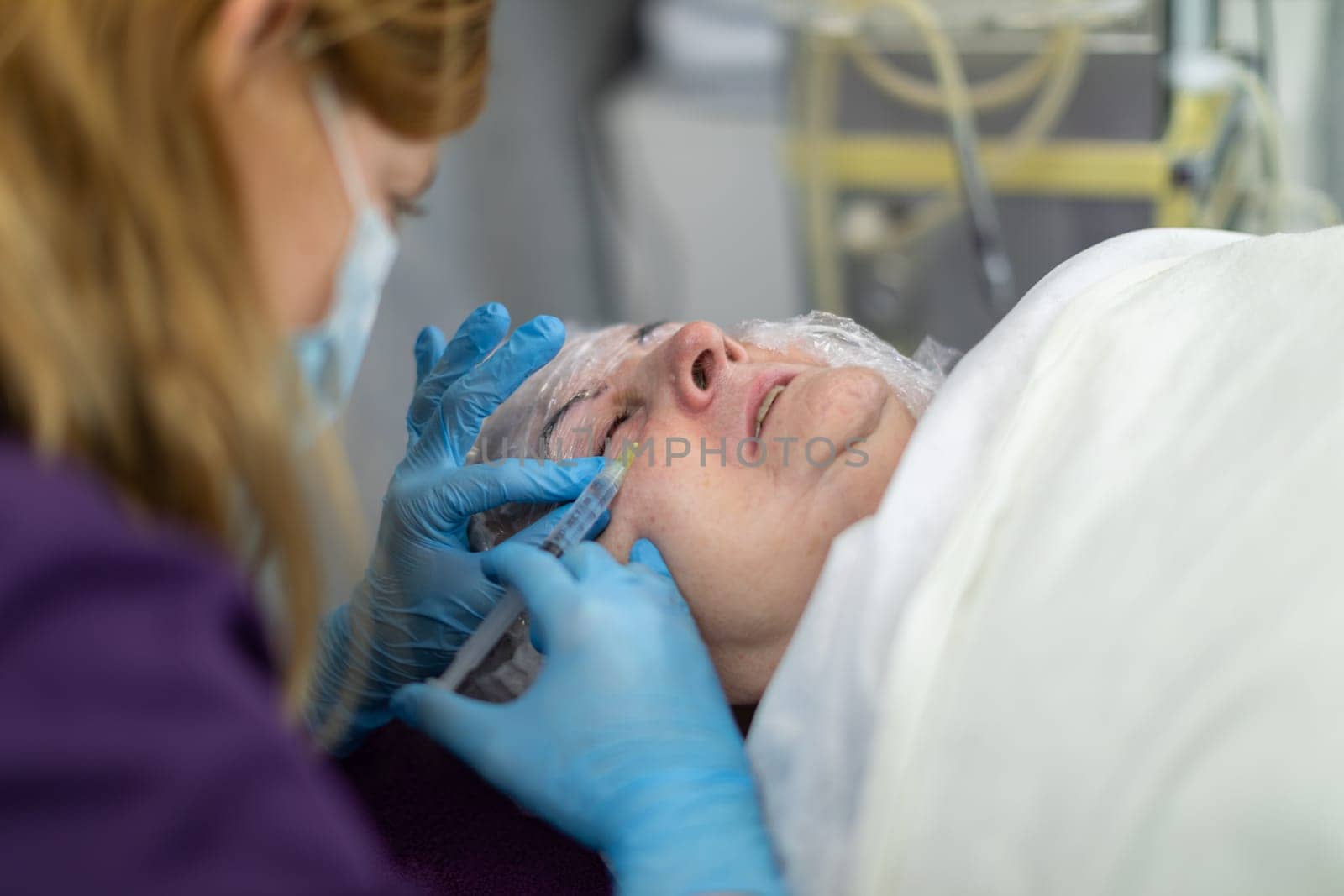 A beauty salon client is lying on a bed. She is having mesotherapy performed. The woman is receiving a series of injections into her face. She contorts her face in a grimace of pain.