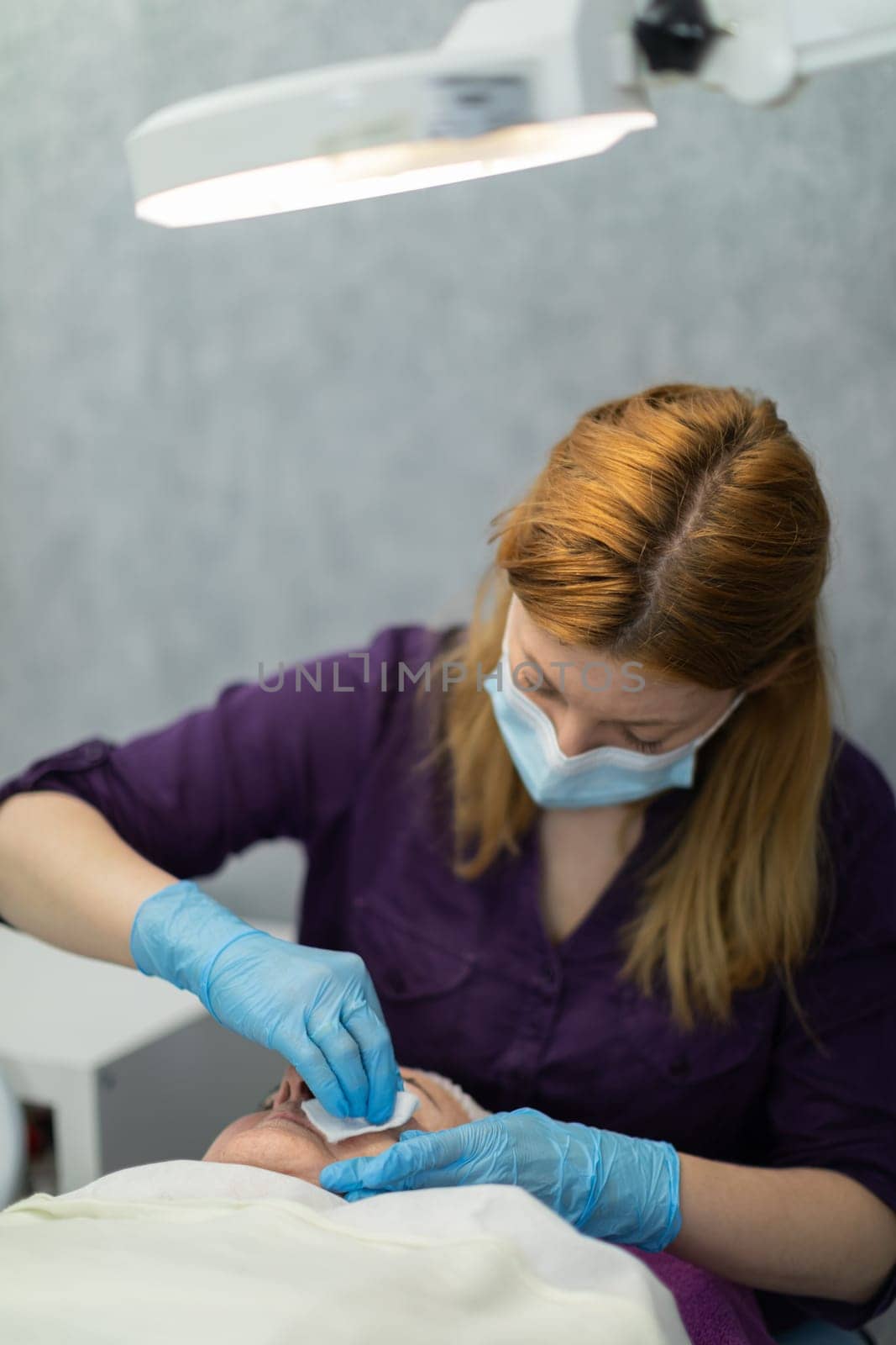 The beautician wipes and disinfects the patient's skin after finishing the mesotherapy treatment. An elderly woman lies on a cosmetic bed while a beautician sits at her head.