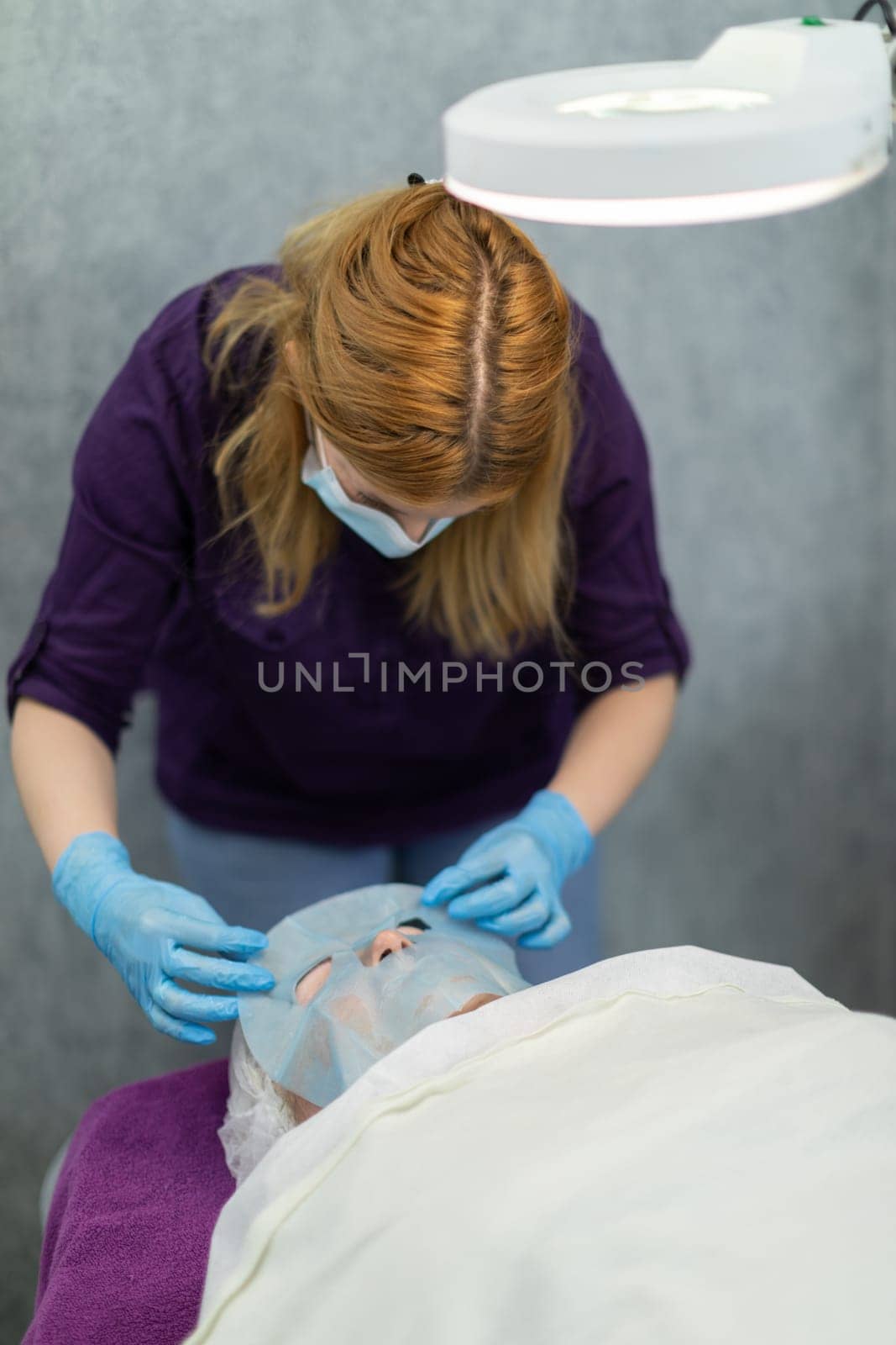 A person making a visit to a beauty salon is lying on a bed. She is covered with a white sheet. The beautician applies a moisturizing mask to the client's face.