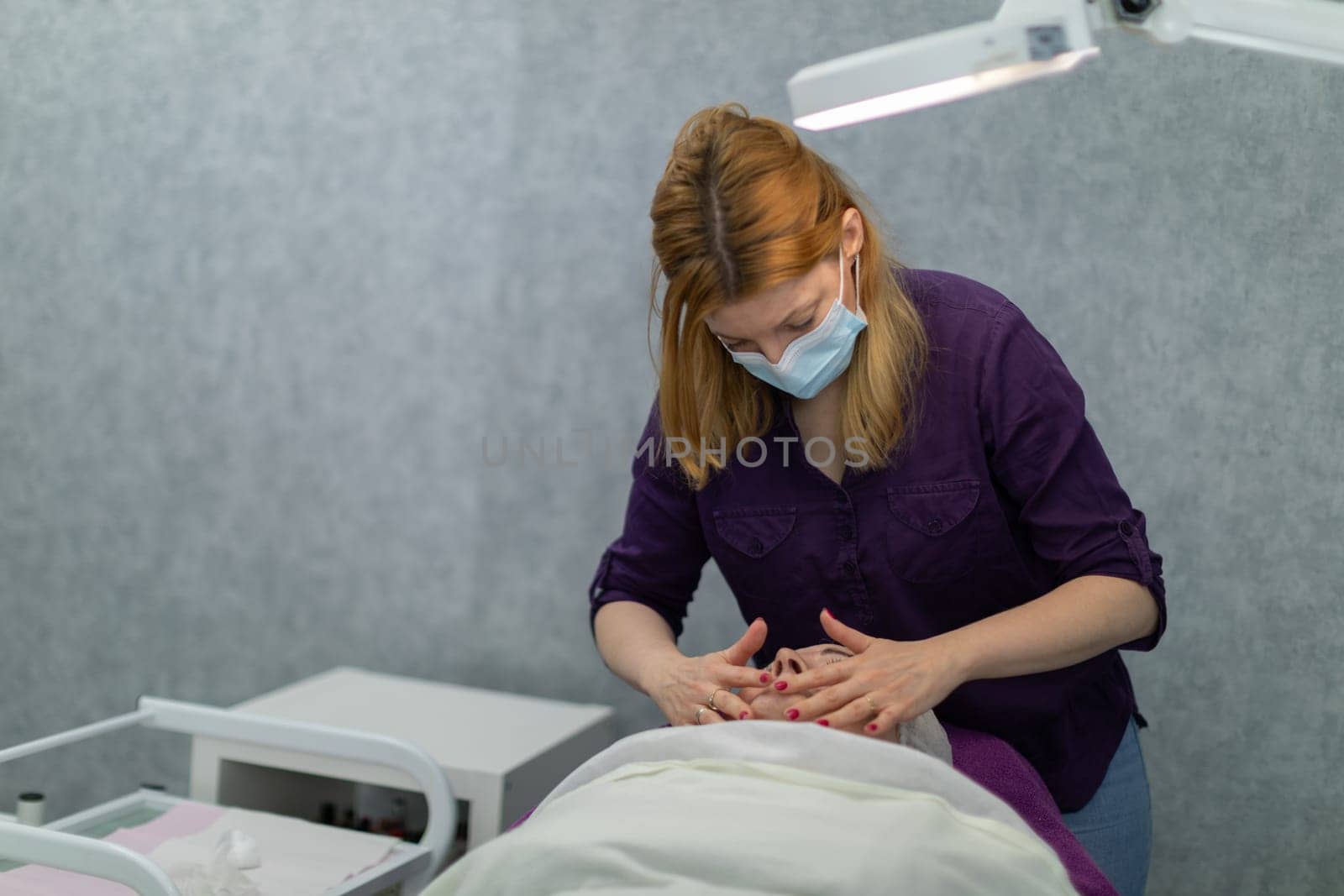 A client of a beauty salon is lying on a bed. The beautician gives her a facial massage. A gray wall can be seen in the background. The patient is illuminated by a medical lamp.