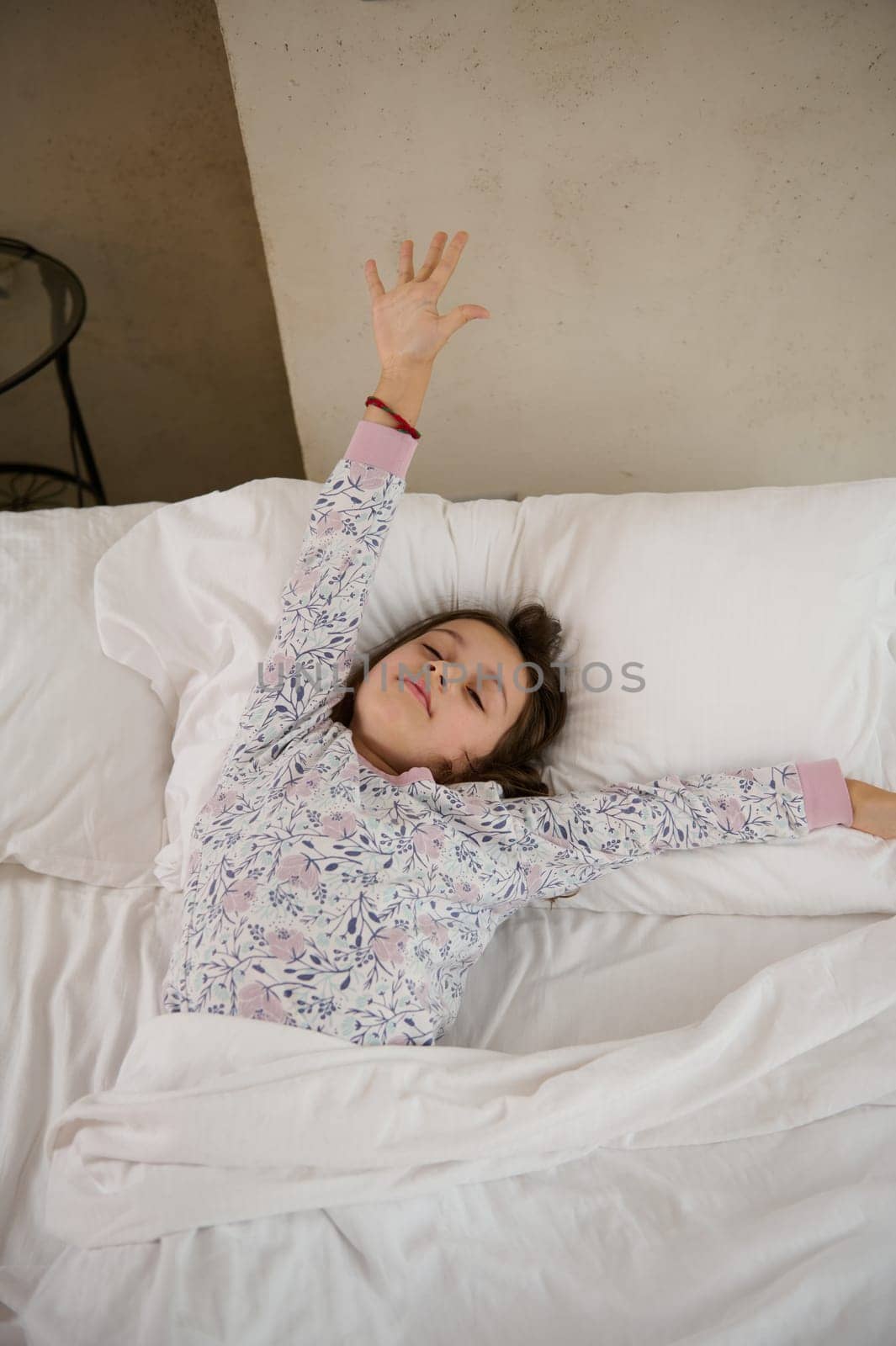 Authentic lifestyle portrait of a happy little child girl lying on the bed and stretching her arms upwards while waking up in the morning