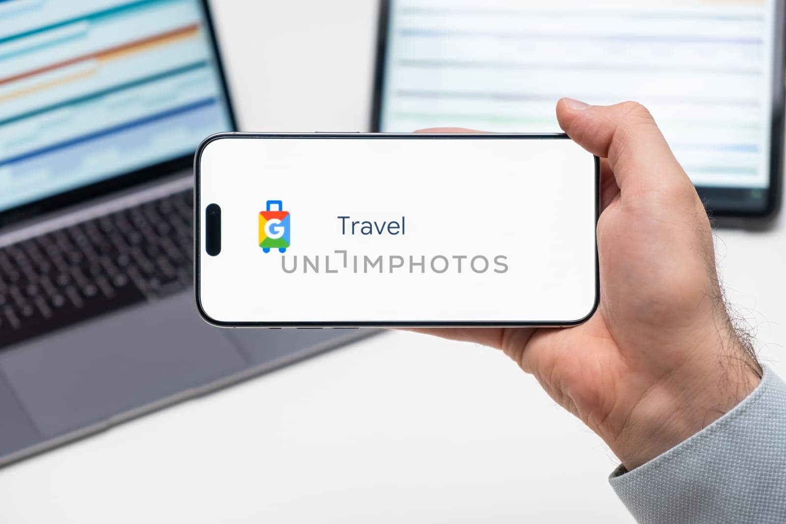 Travel logo of app on the screen of mobile phone held by man in front of the laptop and tablet, December 2023, Prague, Czech Republic