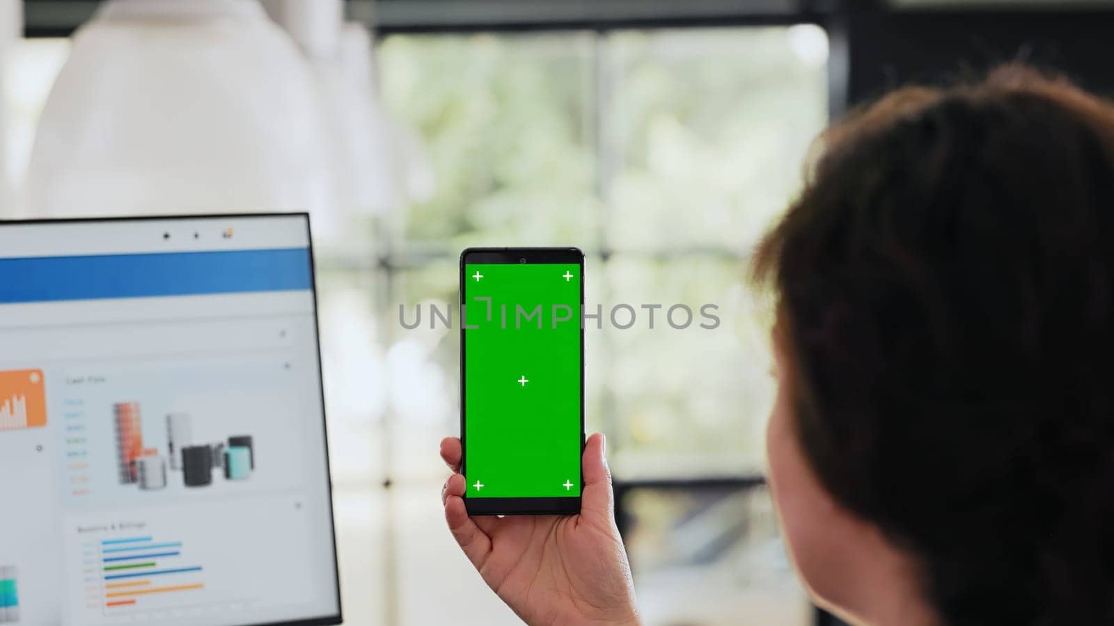 Worker examines phone with greenscreen sitting at workstation in business agency office, analyzing blank chromakey display with copyspace layout. Person works on smartphone. Handheld shot. Close up.