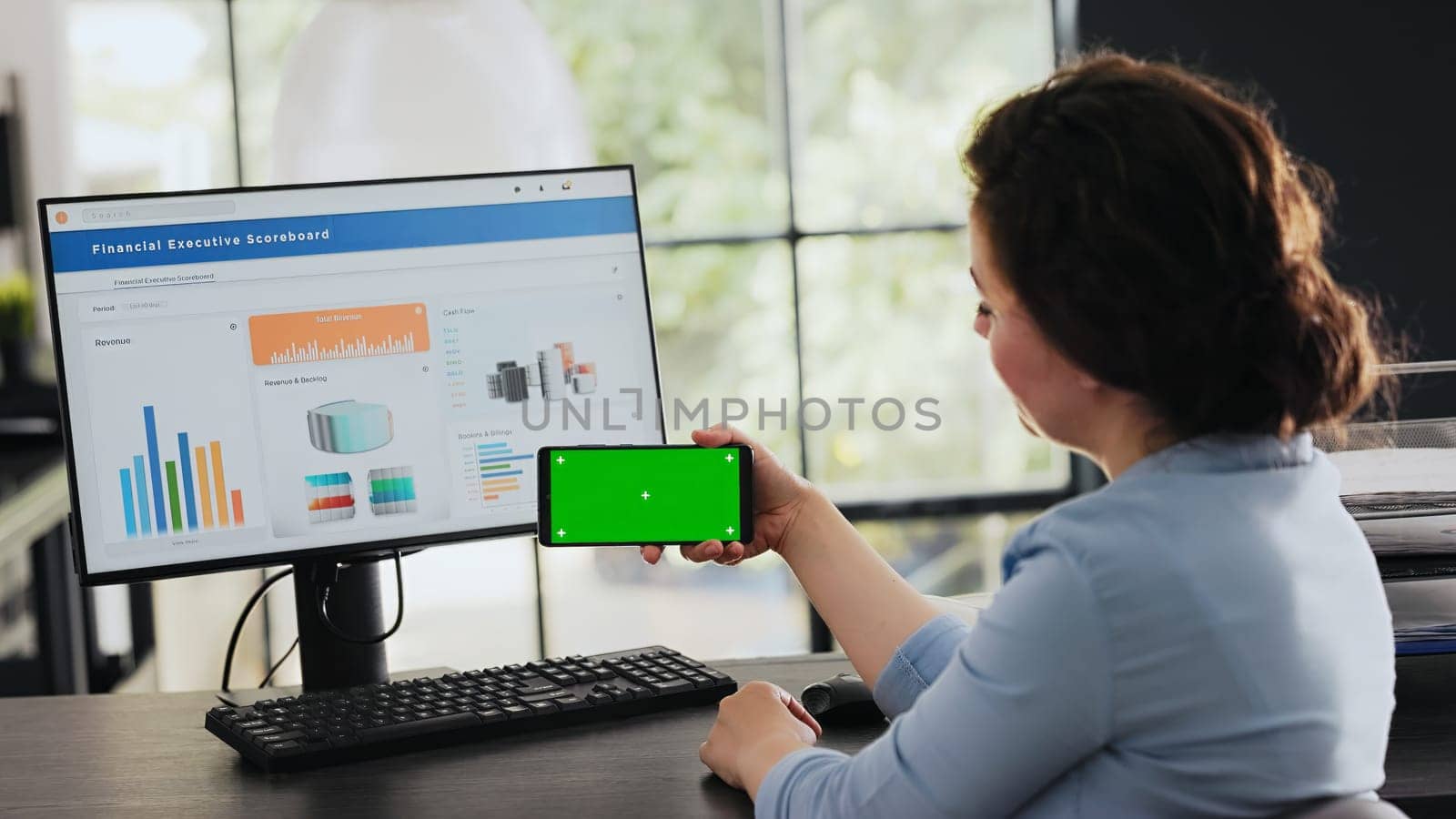 Businesswoman looks at greenscreen while she holds mobile device, isolated chromakey display shown at office desk. Employee checking blank copyspace software on phone. Handheld shot.