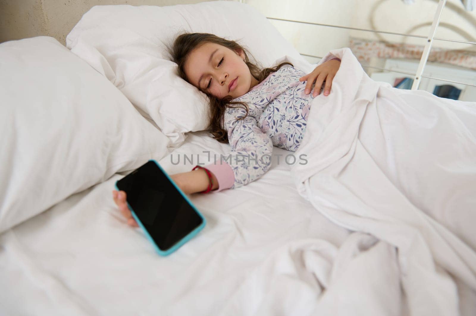 Caucasian child girl in pajamas, falling asleep on her bed with a mobile phone in her hand. Smartphone with black blank mockup digital touch screen. Digital gadget and online video games addiction