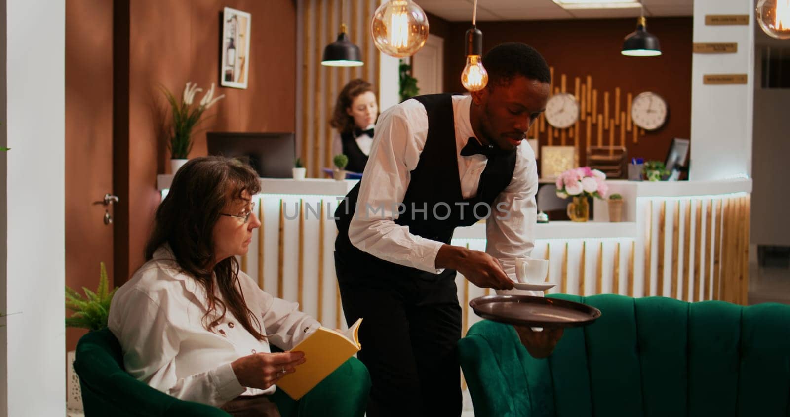 Bellhop serving coffee cup to woman client relaxing in lounge area, senior hotel guest enjoying refreshment and reading book. Employee providing all inclusive services on holiday.