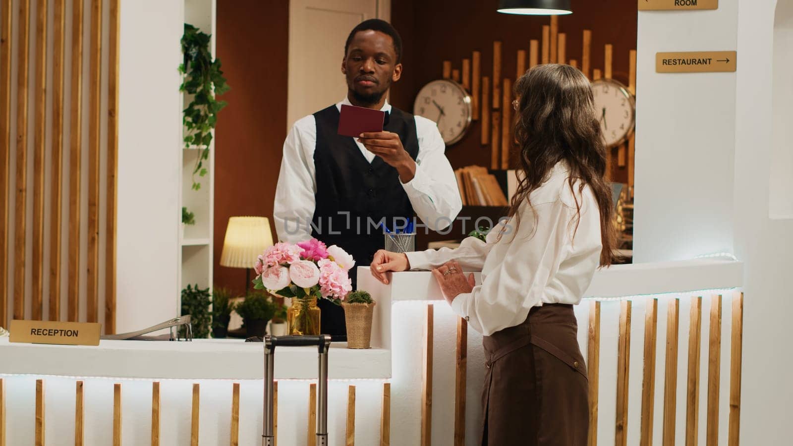 Hotel receptionist assists woman with check in, verifying identity on passport and retrieving reservation details. African american worker checks personal document of tourist at resort.