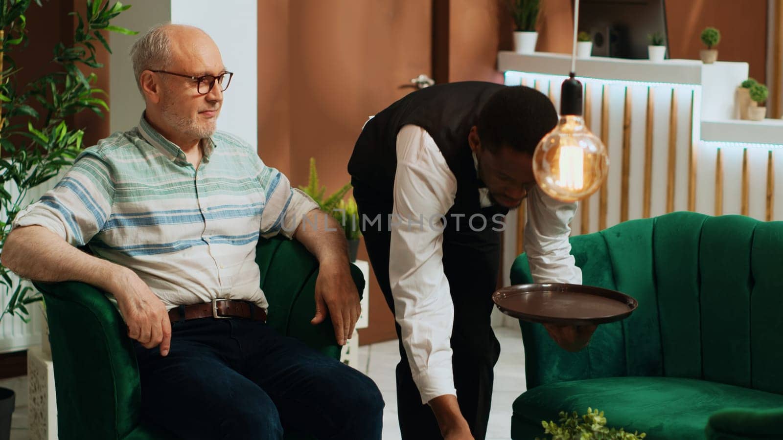 Senior man drinking cup of coffee in lounge area and waiting for room reservation, african american bellhop serving hotel guest with refreshment from bar. Elderly person receiving good staff service.