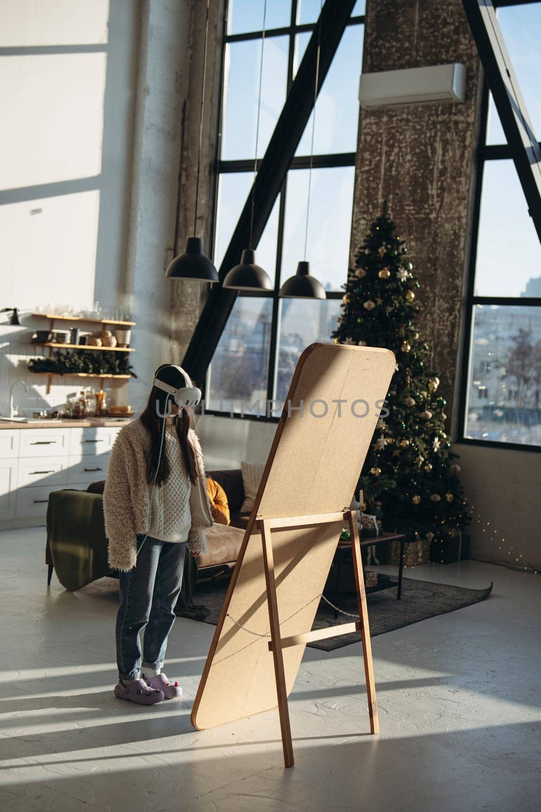 A young woman in light clothing and a virtual reality headset stands in front of the mirror in a Christmas atmosphere. High quality photo