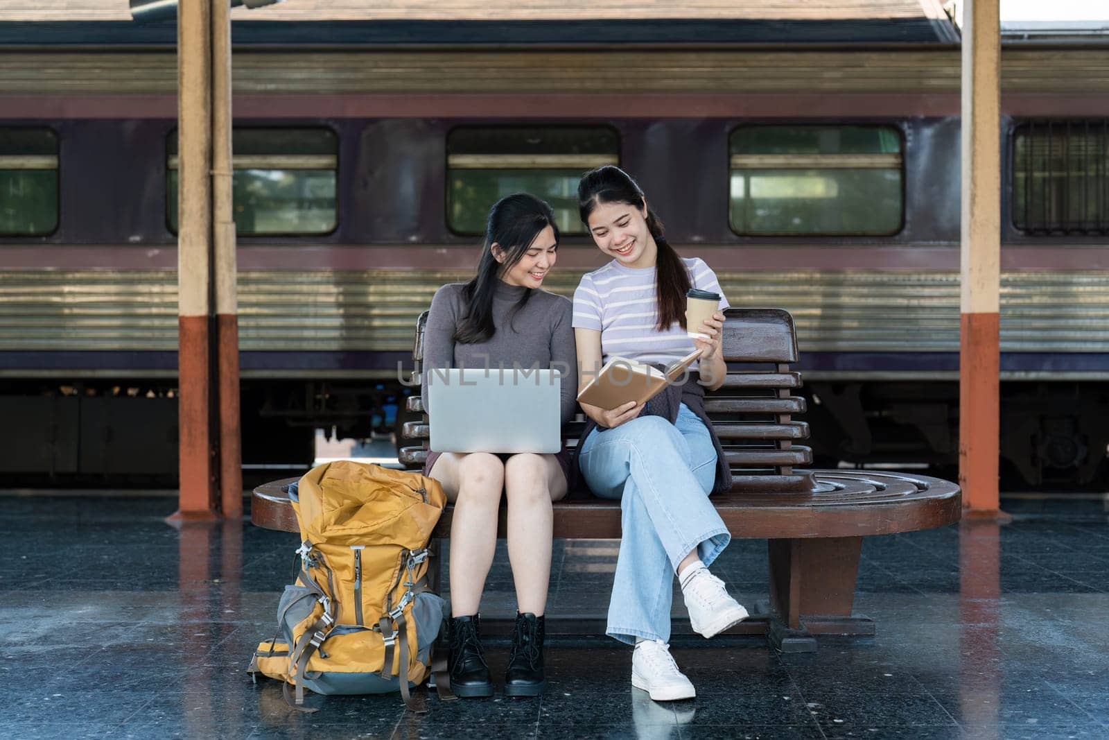 Asian woman friends using laptop and note for planing trip together at railway station have happy moment.