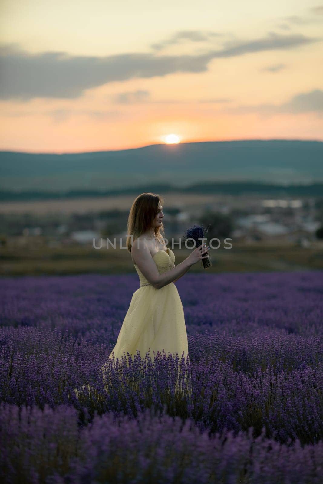 Woman poses in lavender field at sunset. Happy woman in yellow dress holds lavender bouquet. Aromatherapy concept, lavender oil, photo session in lavender by Matiunina