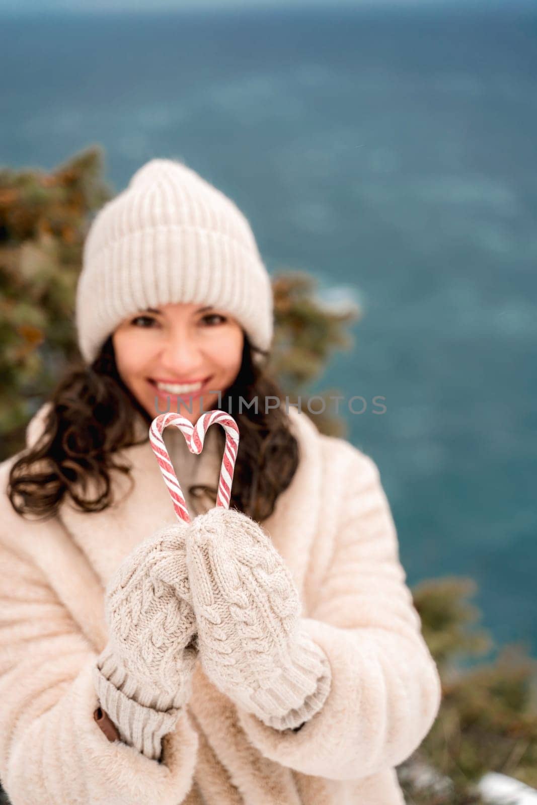 Woman candy sea. Smiling woman in knitted hat, mittens and beige coat holding lollipops candy canes in her hands in shape of heart against the backdrop of the sea