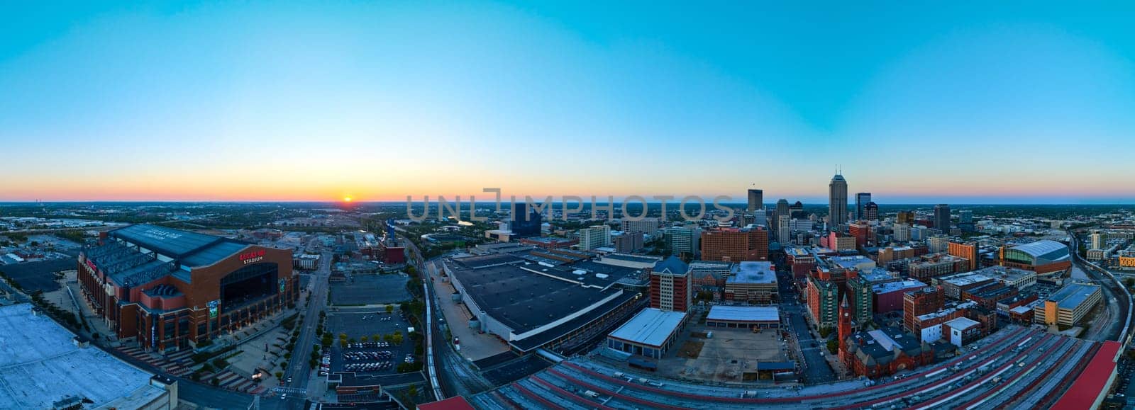 Dusk descends over Indianapolis, showcasing an illuminated Lucas Oil Stadium and vibrant skyline in a serene panorama, captured from an aerial perspective.