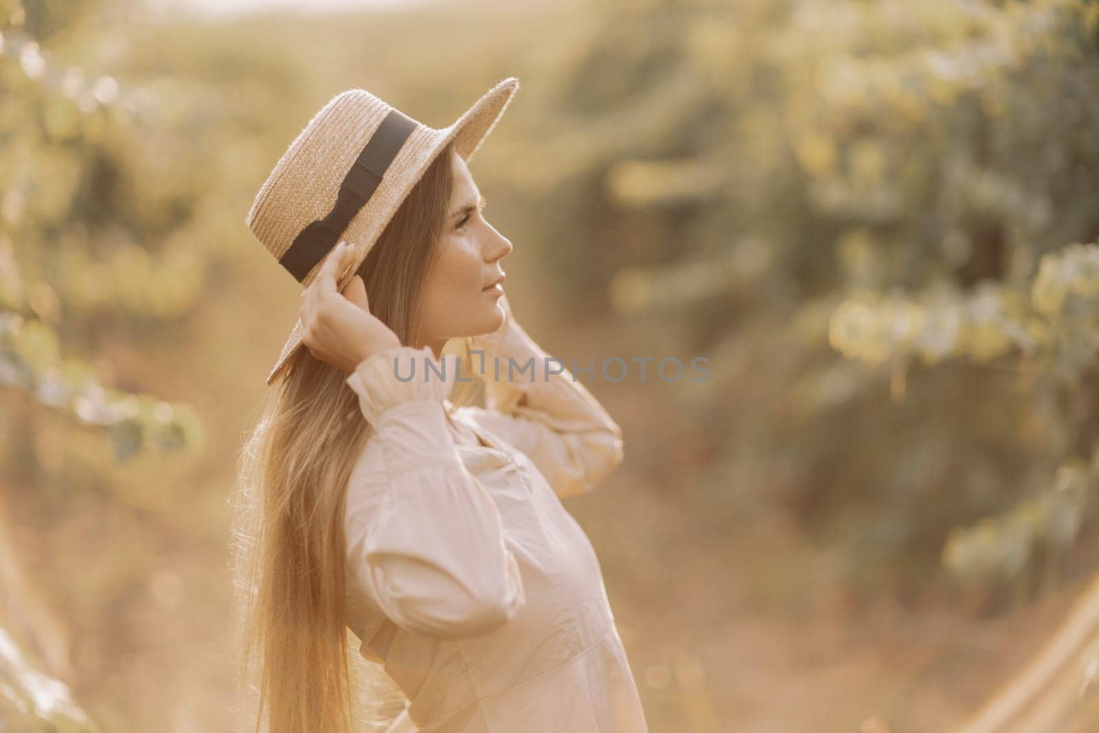 Woman with straw hat stands in front of vineyard. She is wearing a light dress and posing for a photo. Travel concept to different countries.