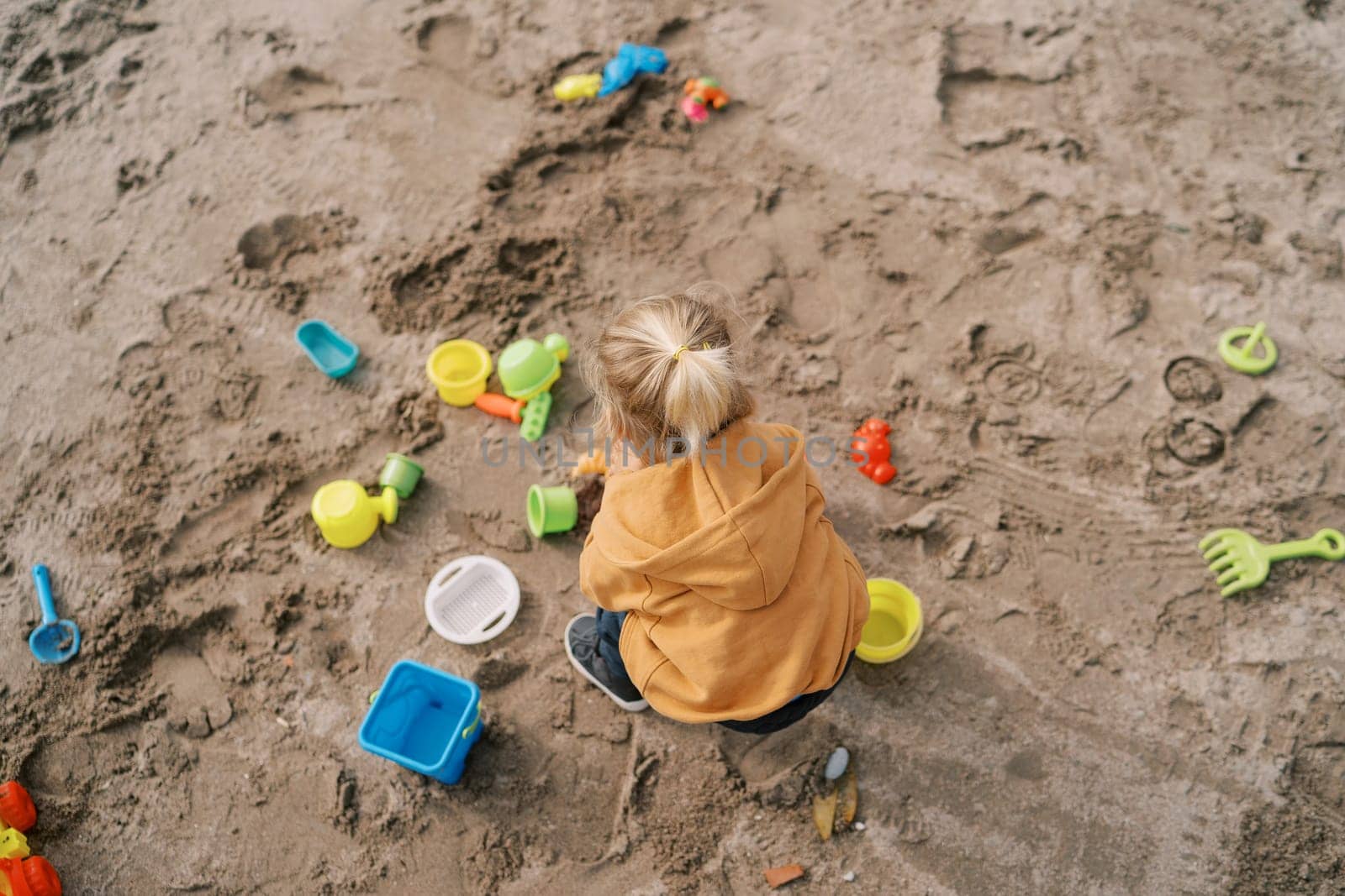 Little girl squats on the sand and plays with colorful toys. Top view by Nadtochiy