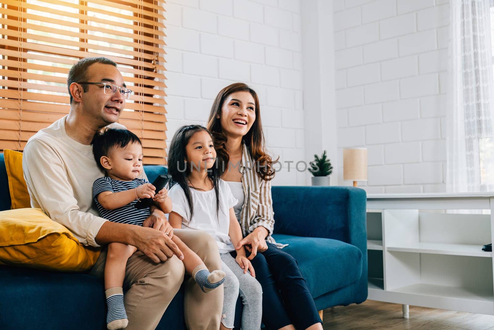 A happy family with kids finds relaxation and togetherness while watching TV movies on the sofa in their grooved modern living room. The father mother son and daughter share smiles laughter and fun.
